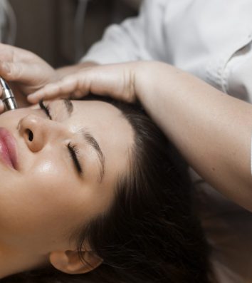 Close up side view portrait of a cute caucasian woman doing noninvasive microdermabrasion on her face with a dermapen by a cosmetician.